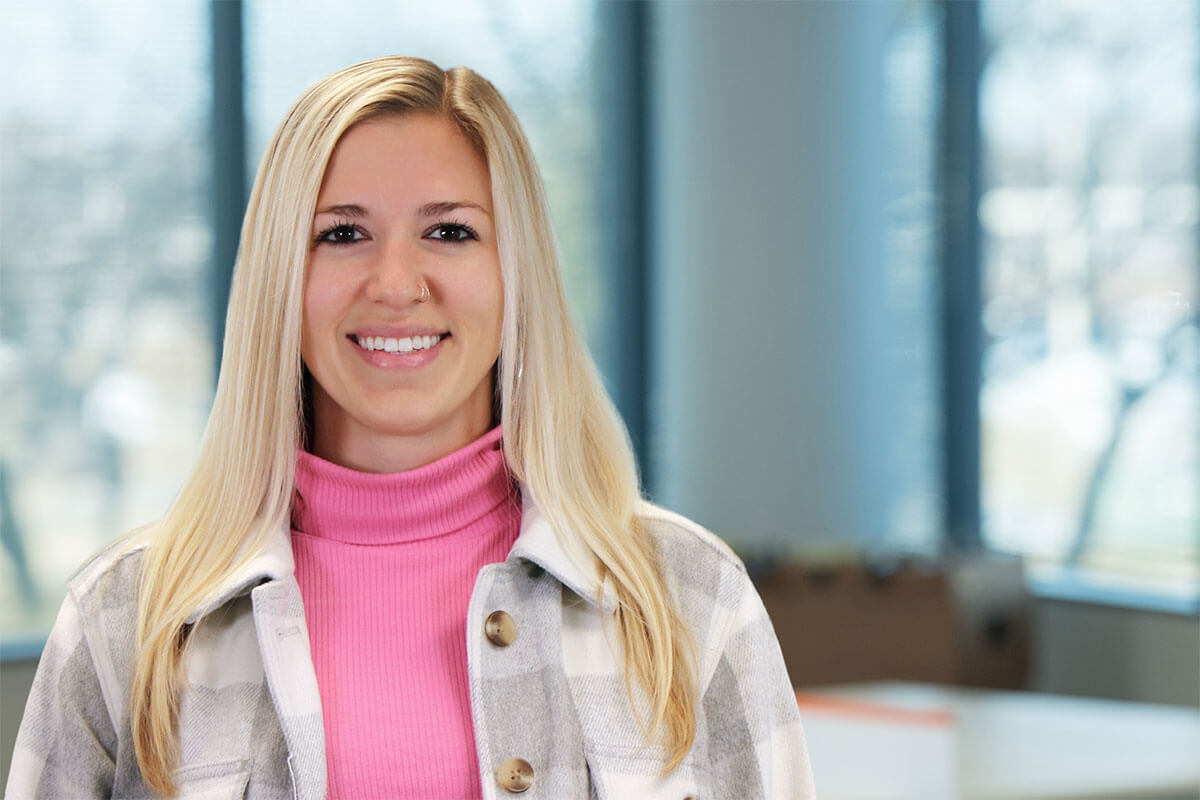 Smiling blonde woman wearing a pink turtleneck and plaid jacket stands in a bright office with large windows and a modern workspace in the background.