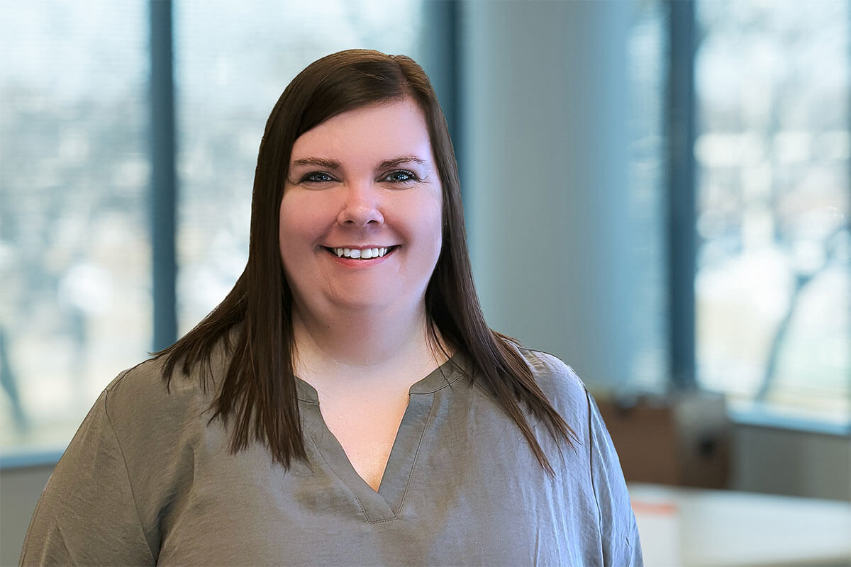 Smiling brunette woman wearing a gray blouse stands in a bright office with large windows and a softly blurred workspace in the background.