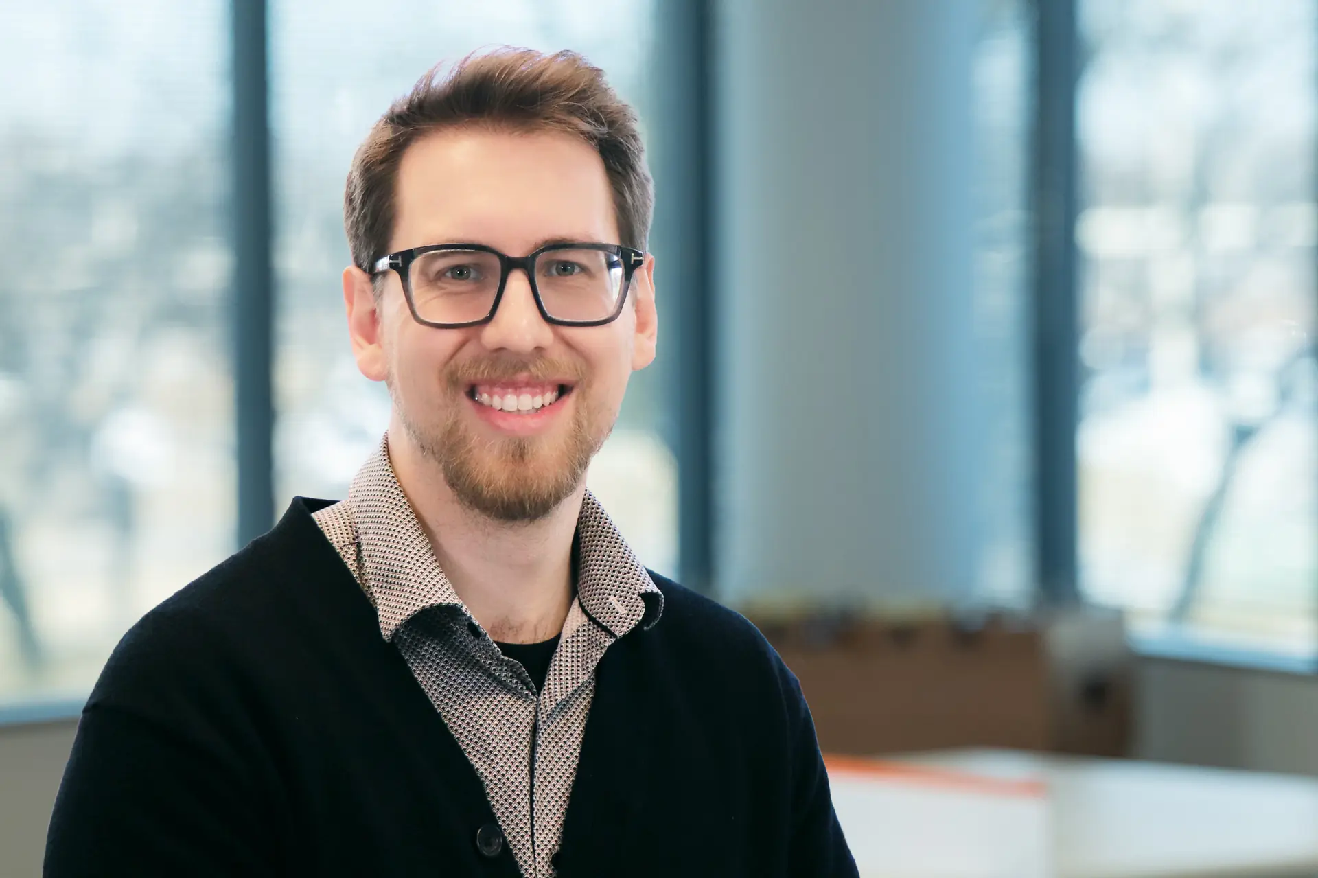 Smiling man with glasses and short brown hair wearing dark cardigan in office setting