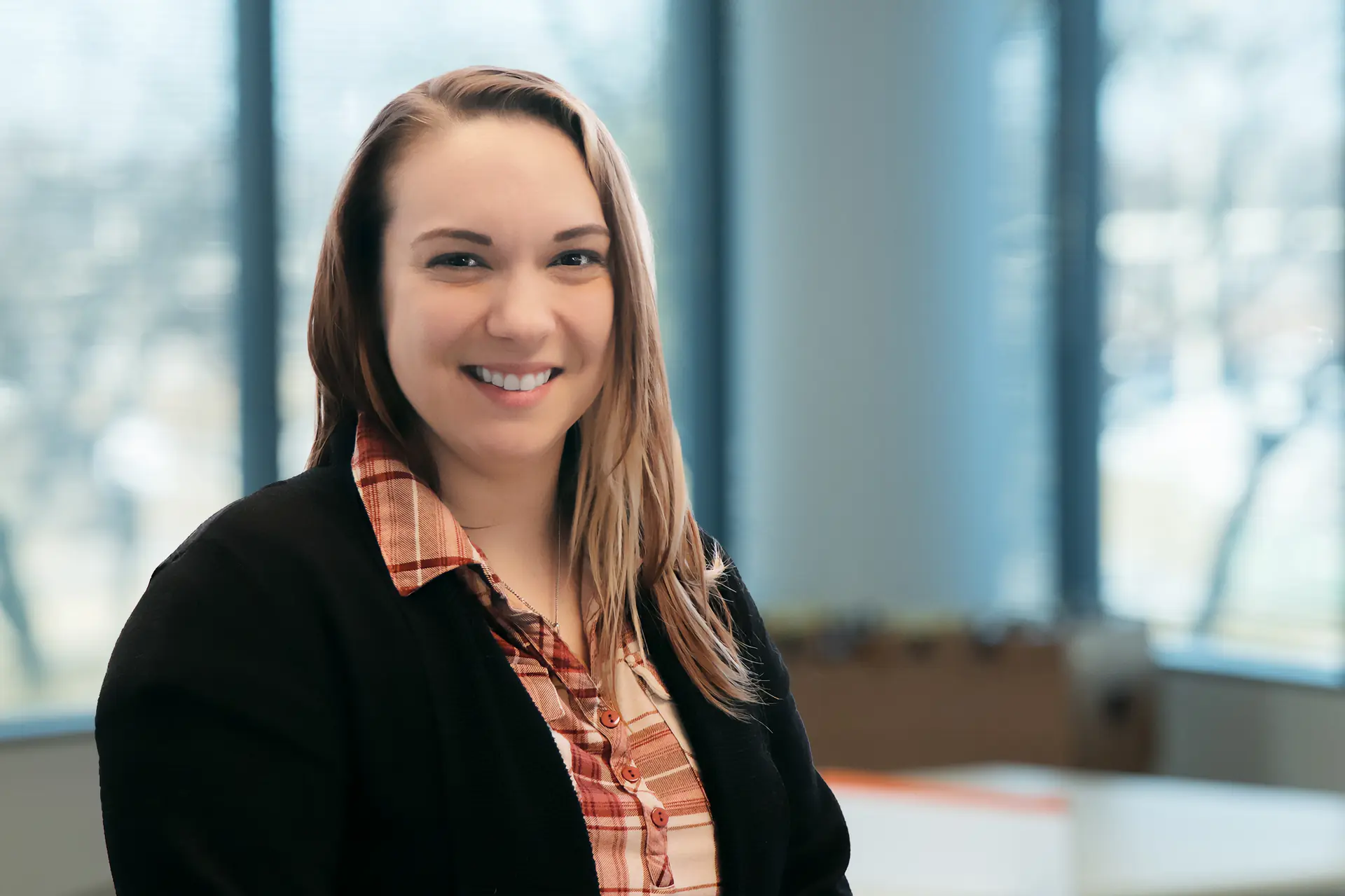 Smiling woman with long light brown hair wearing plaid shirt and black blazer in office