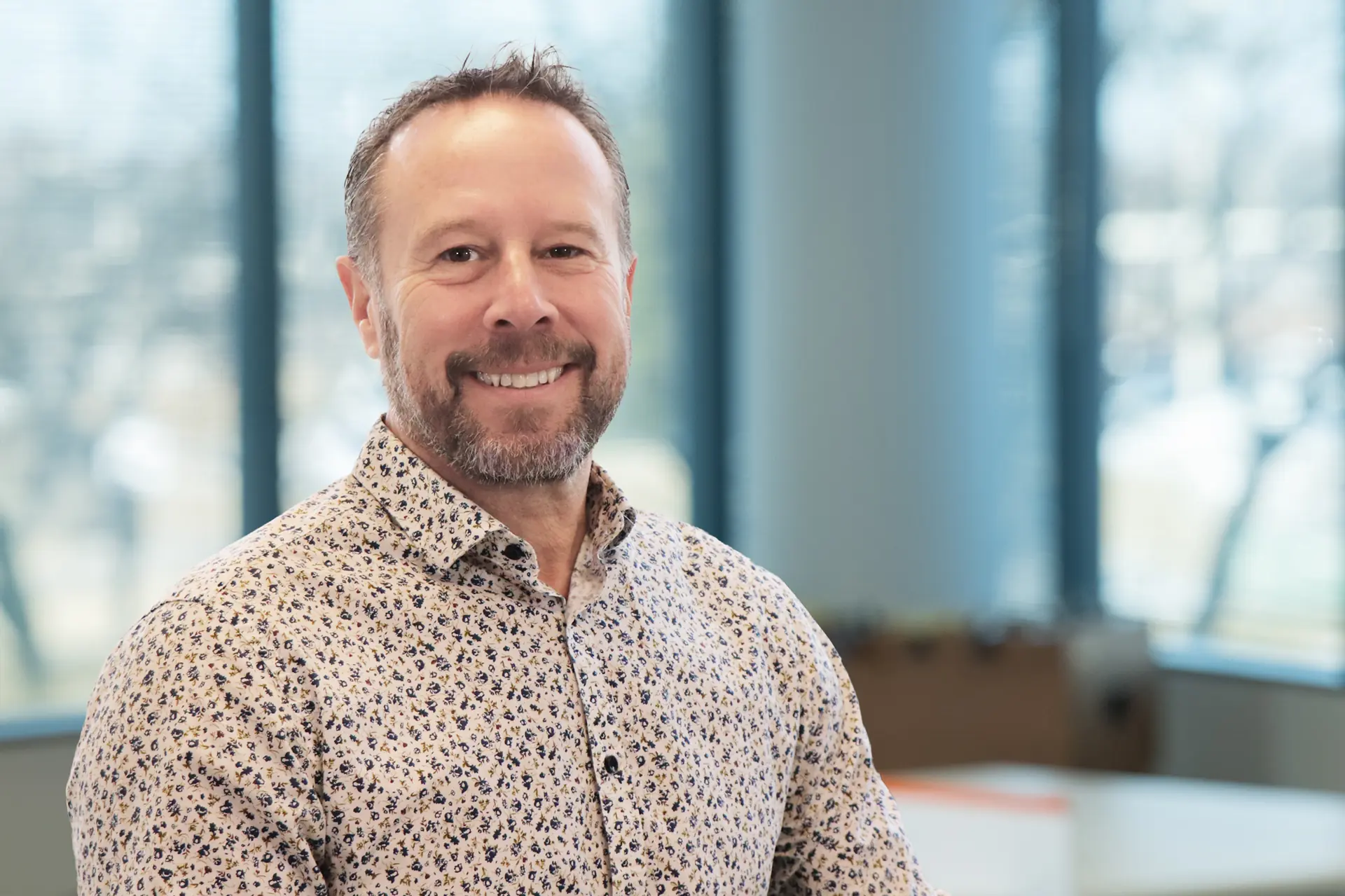Smiling man with short hair and trimmed beard wearing patterned shirt in office setting